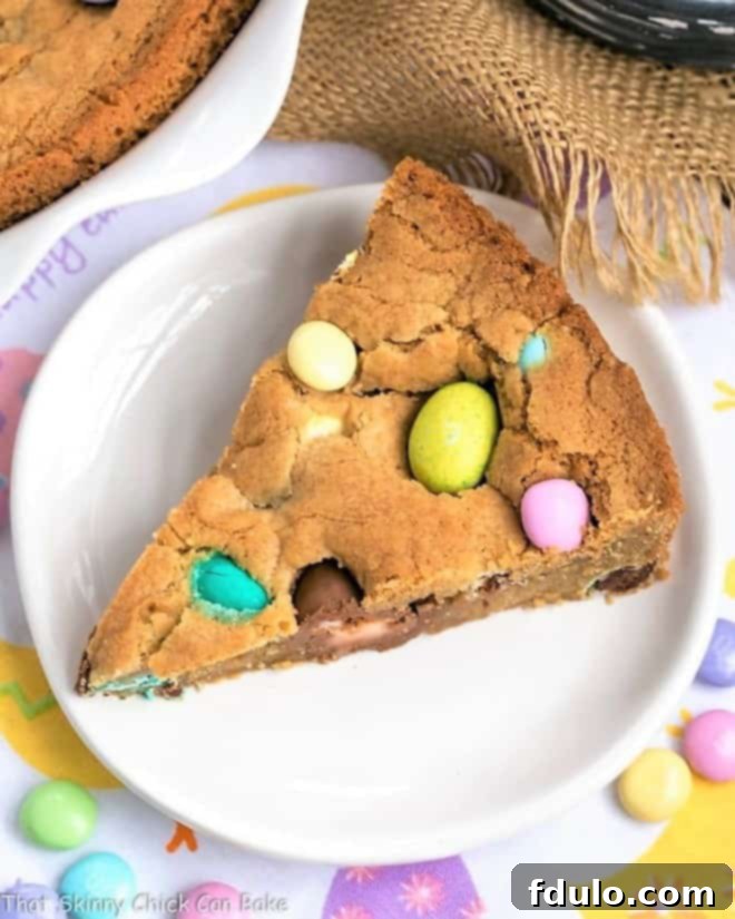 Homemade Cookie Cake - A giant, chewy cookie packed full of chocolate chips and Easter candy A close-up of a generous slice of homemade Easter cookie cake, showcasing its thick, chewy texture, abundant chocolate chips, and colorful Easter candy inclusions, presented on a simple white dessert plate.