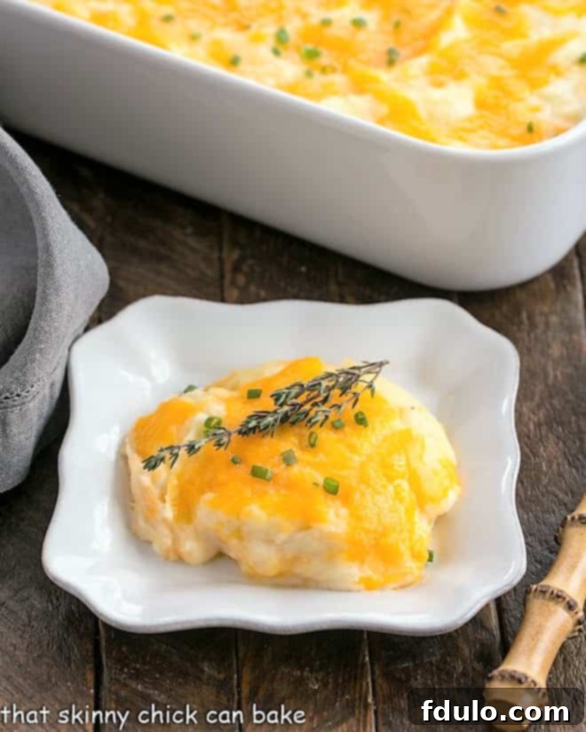 A serving of creamy cheesy mashed potato casserole on a white square plate with the full baking dish in the background, garnished with fresh herbs.