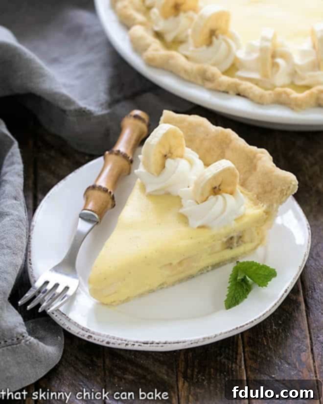 A perfectly sliced piece of banana cream pie rests on a white plate with a bamboo-handled fork, with the full pie subtly blurred in the background, highlighting its delicious layers.