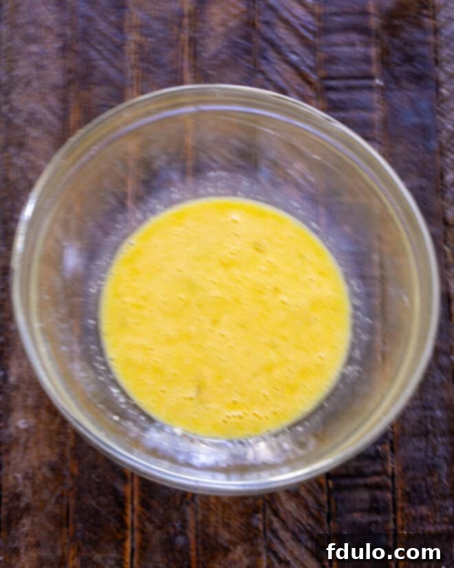 A close-up shot of two slightly beaten eggs, swirled gently in a clear glass bowl, with a generous sprinkle of grated Parmesan cheese mixed in, forming a creamy golden liquid ready for the next step of the recipe.