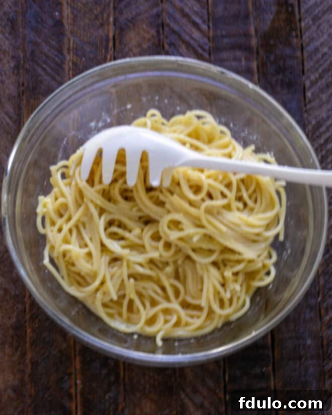 Cooked spaghetti strands, perfectly separated and glistening, are carefully tossed with the egg and Parmesan mixture in a large mixing bowl, ensuring an even coating on every single strand.