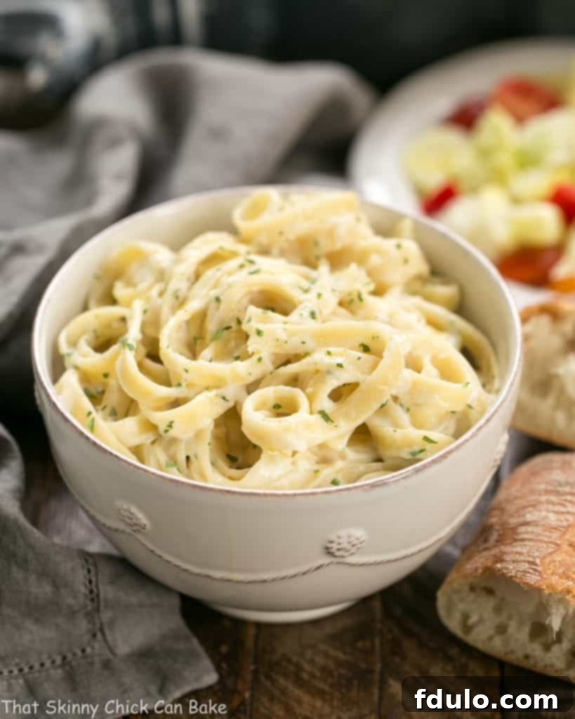Light Fettuccine Alfredo in a white bowl, with crusty bread and a fresh salad blurred in the background, suggesting a complete meal.