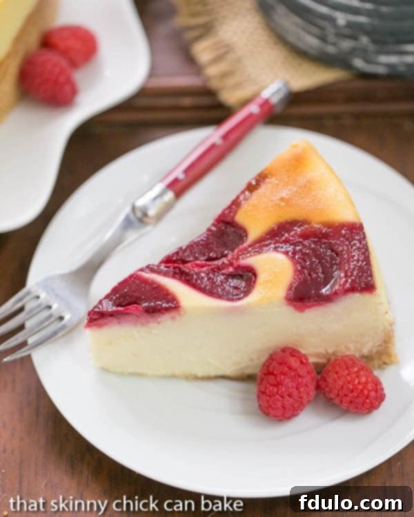A close-up of a White Chocolate Raspberry Swirl Cheesecake slice on a white plate with a red handled fork, highlighting its creamy texture.
