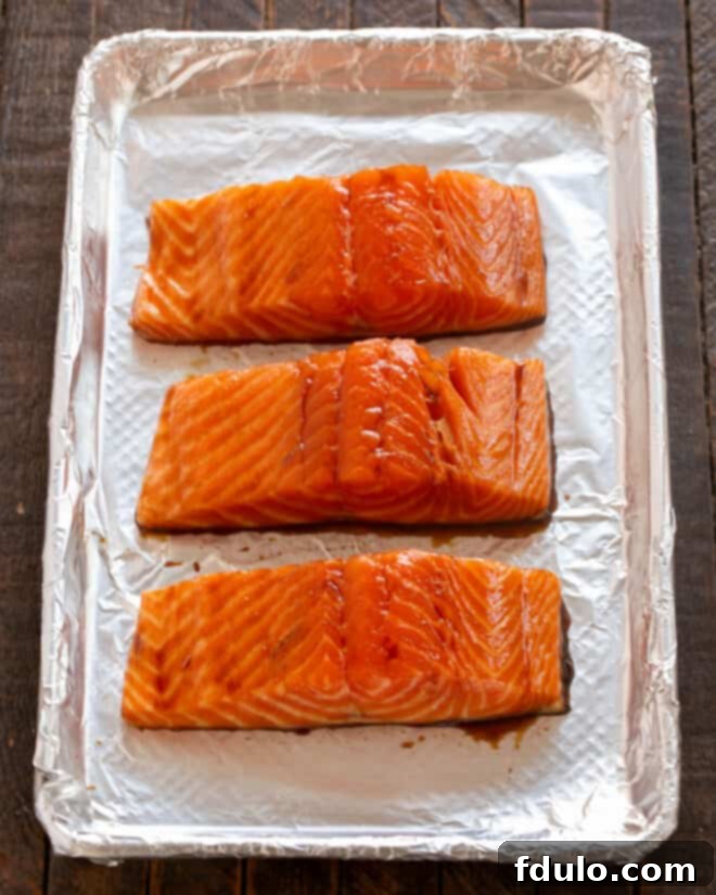 Marinated salmon fillets arranged on a sheet pan, ready for broiling. A brush is poised to apply additional marinade.