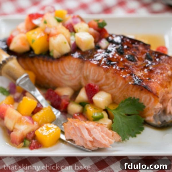 A serving of Maple Glazed Salmon on a white plate, with a fork holding a flaky piece of salmon next to a larger fillet, showcasing its texture and the vibrant salsa.