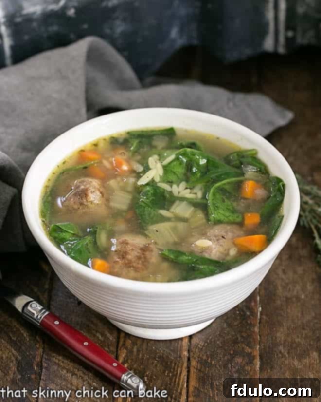 A generously filled white bowl of Italian Wedding Soup, featuring small meatballs, green spinach, and tiny pasta, served with a red-handled spoon.