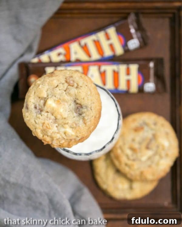 A tempting stack of golden brown White Chocolate Toffee Chunk Cookies, showcasing their delightful texture, positioned next to a tall glass of milk, garnished with pieces of Heath bars and a rustic gray napkin.