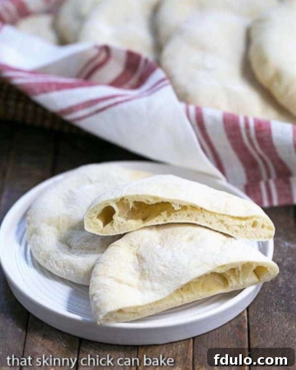 Freshly baked homemade pita bread on a white ceramic plate, with one pita sliced in half to reveal its pocket, showcasing its fluffy texture.