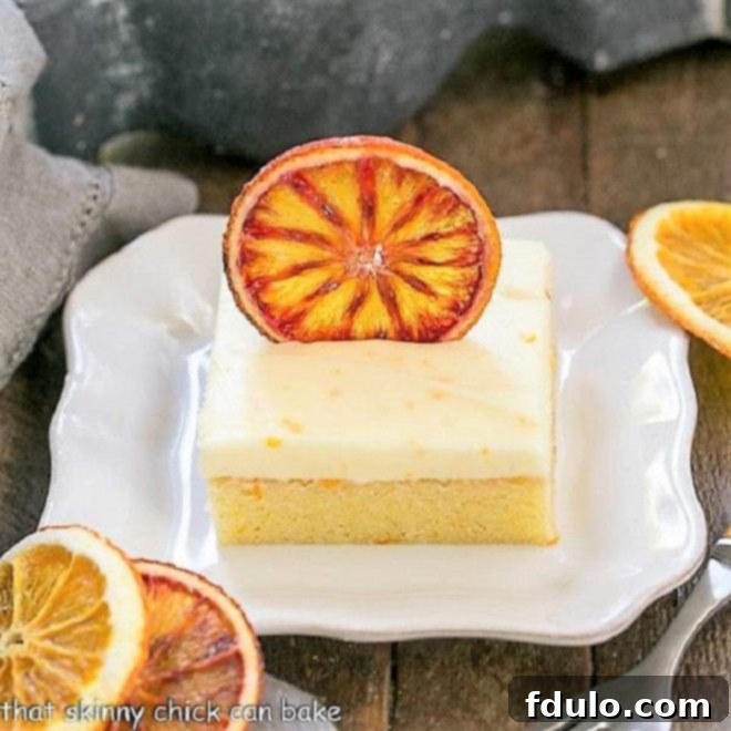 Close-up of a slice of dense Orange Cake with thick Cream Cheese Frosting, garnished with dried orange slices on a white plate.