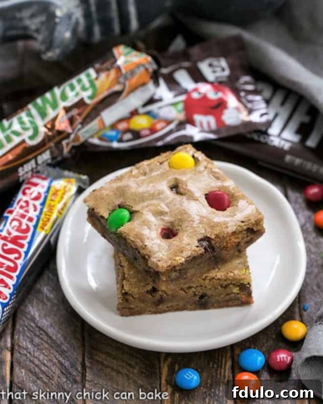 Candy Bar Blondies - The perfect way to use up any leftover Halloween candy! Two golden Candy Bar Blondies, generously studded with colorful candies and chocolate, stacked appealingly on a small white plate.