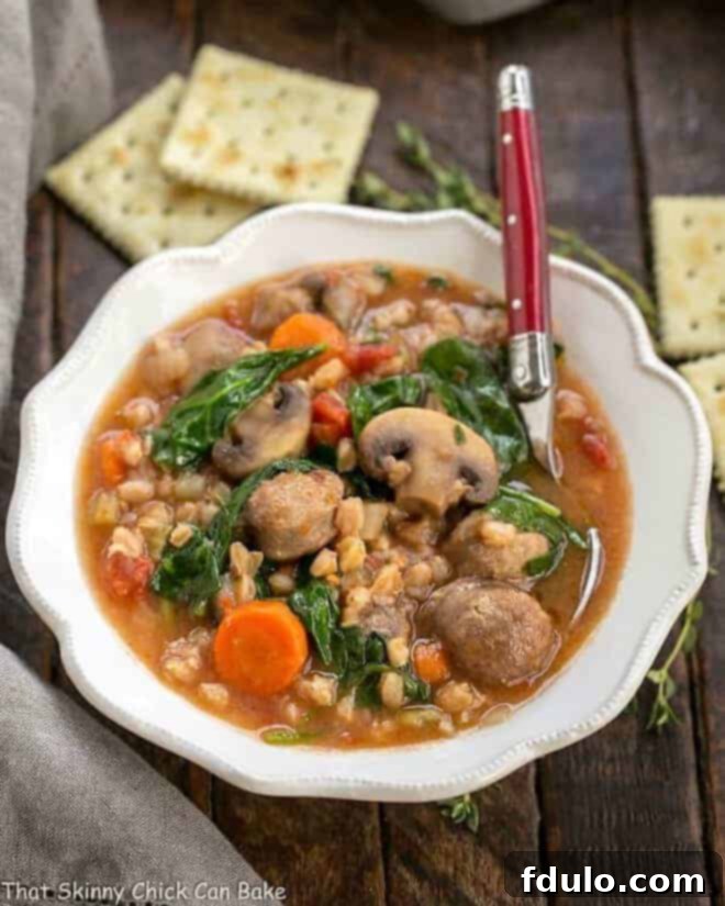 A close-up of Rustic Farro Soup in a beautifully scalloped white soup bowl, with a silver spoon poised to scoop a delicious bite.