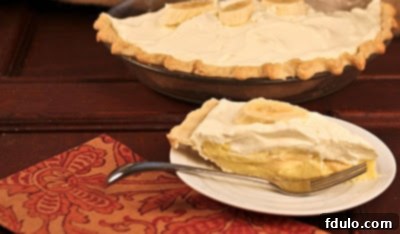 A generously sized slice of Banana Cream Pie served on a dessert plate, with the remainder of the pie elegantly blurred in the background.