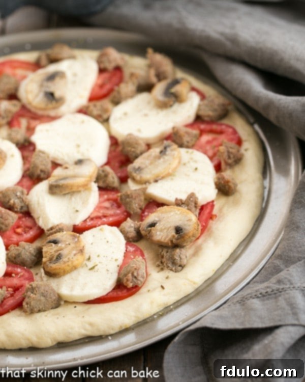 Homemade Pizza Dough crafted with yeast, generously topped with creamy mozzarella, juicy tomatoes, savory sausage, and earthy mushrooms, waiting to be transformed in the oven! Sausage Mushroom Pizza on a pizza pan, perfectly topped and ready to go into the hot oven for baking.
