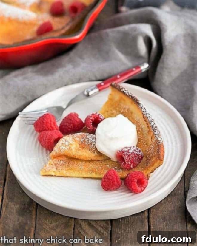 A golden-brown Dutch Baby Pancake slice on a white plate, elegantly topped with fresh raspberries, a dusting of powdered sugar, and a dollop of sweetened whipped cream.