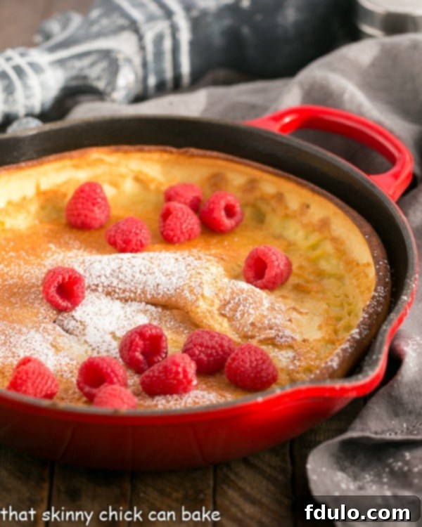 A vibrant red cast iron skillet filled with a freshly baked Dutch Baby Pancake, generously adorned with bright red raspberries and a delicate dusting of powdered sugar.