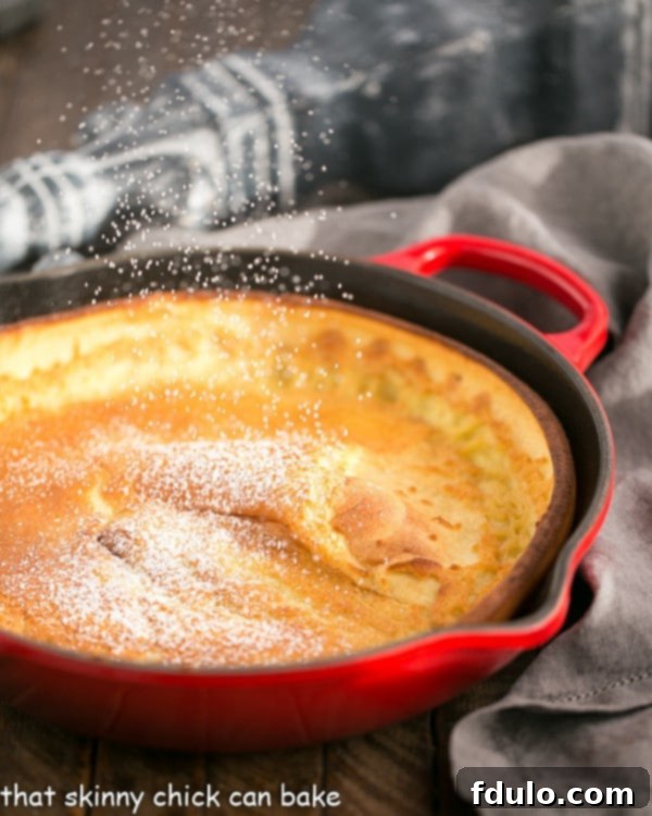 A close-up shot of a beautifully puffed Dutch Baby Pancake in a red cast iron skillet, lightly dusted with powdered sugar, showcasing its golden-brown, crispy edges.
