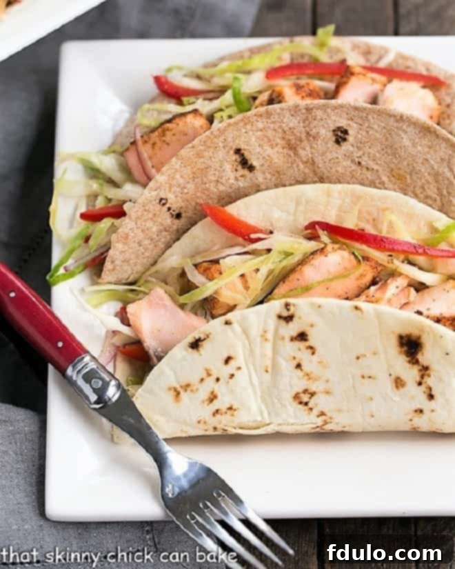 Salmon Tacos with Mango Salsa - Roasted salmon with all the fixings for a sophisticated seafood taco Two Salmon Tacos with vibrant Mango Salsa and creamy Sriracha Lime Sauce, served on a square white plate with a red handled fork, showcasing a gourmet and healthy fish taco experience.