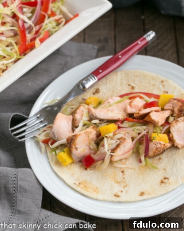 Fish Tacos Recipe with Salmon and Mango Salsa - Roasted salmon with all the fixings for a sophisticated seafood taco Close-up of a single Salmon Taco, garnished with fresh Mango Salsa and vibrant Cabbage Slaw, artfully arranged on a round white plate with a red handled fork, emphasizing the rich textures and colors.