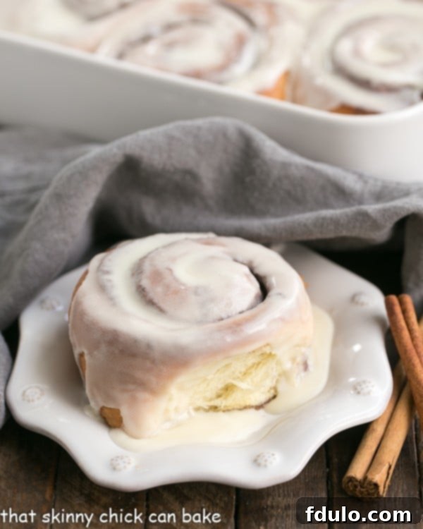 A freshly baked cinnamon bun on a white plate, with a pan full of other glazed buns blurred in the background, highlighting their readiness.