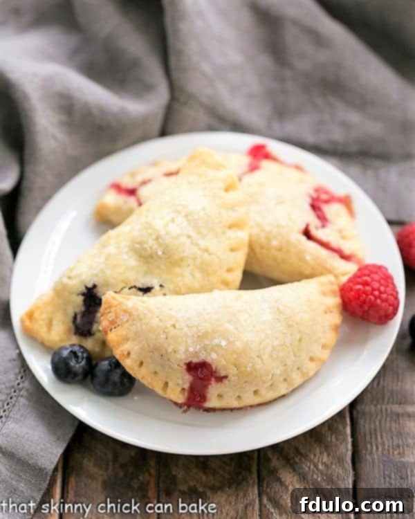 Rustic Berry Turnovers - Buttery crusts + sweetened berries make these fabulous hand pies! Several Rustic Berry Turnovers arranged on a white serving plate, showcasing their golden crusts and slight berry ooze.
