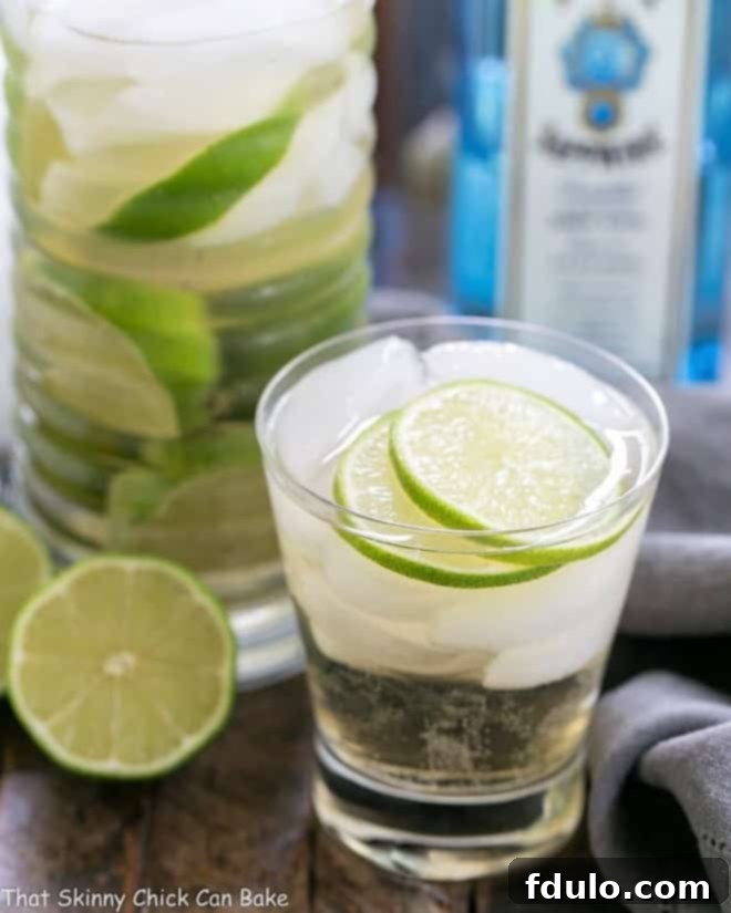 Close-up of a St. Germain Gin and Tonic Cocktail in a short glass, brimming with ice cubes and adorned with multiple lime slices, on a blurred background.