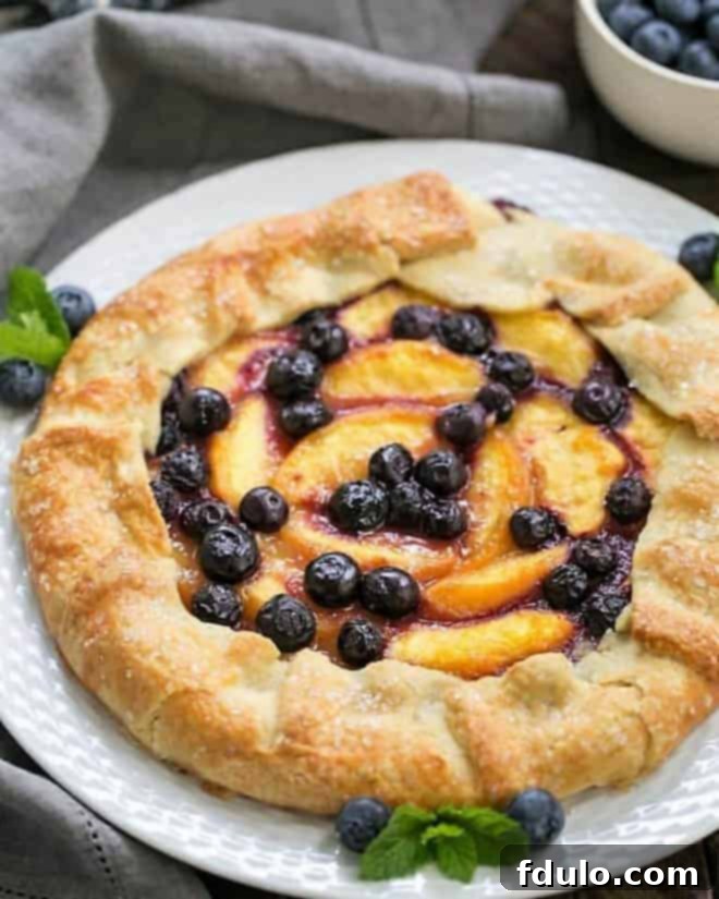 Peach Blueberry Galette - A gorgeous summer dessert with all the flavors of pie without all the work! A beautifully baked Peach Blueberry Galette on a white serving plate, showcasing its golden, flaky crust and vibrant peach and blueberry filling.
