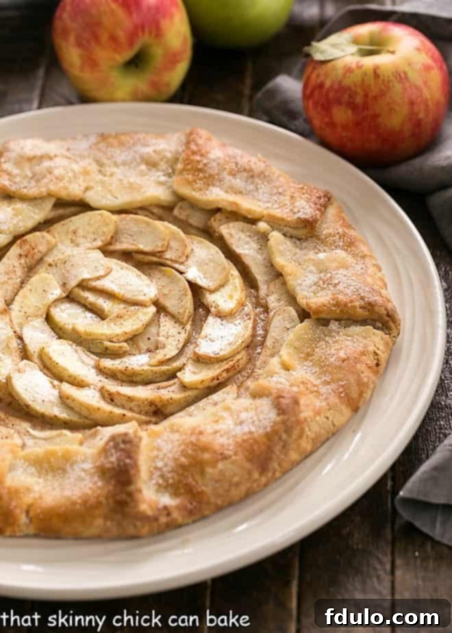 A slice of Rustic Apple Tart on a white plate, with whole apples blurred in the background, highlighting the dessert's delicious appearance.