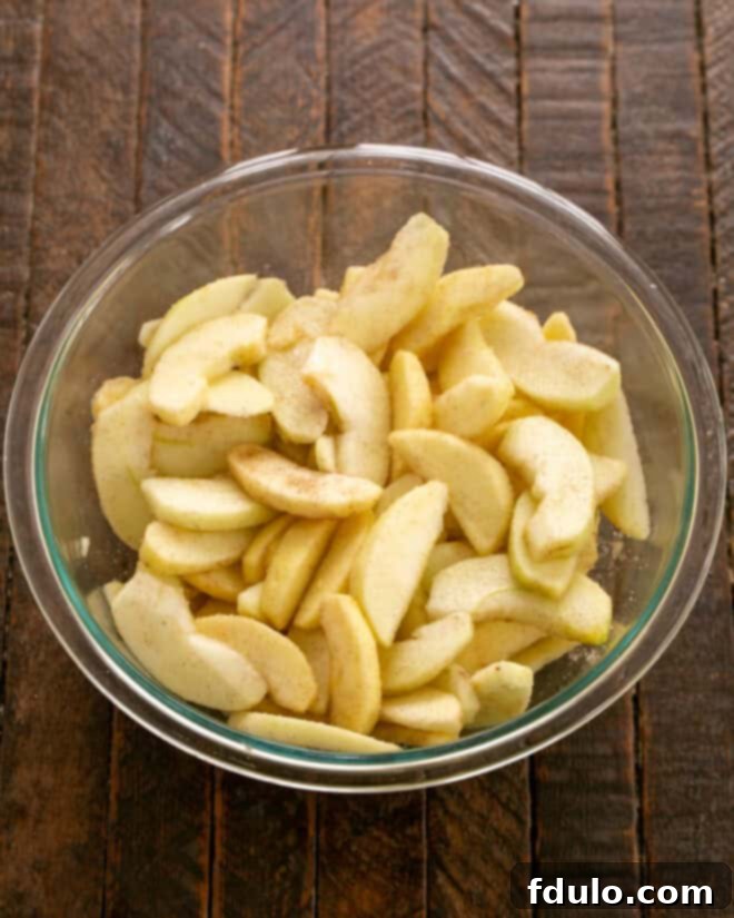 Peeled, thinly sliced apples tossed with cinnamon, sugar, and lemon juice in a clear glass bowl, preparing them for the galette filling.