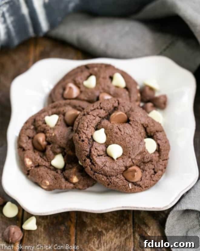 Close-up of freshly baked Triple Chocolate Fudge Cookies, featuring a rich, dark cocoa base studded generously with melted milk and white chocolate chips, resting on a pristine white square plate.