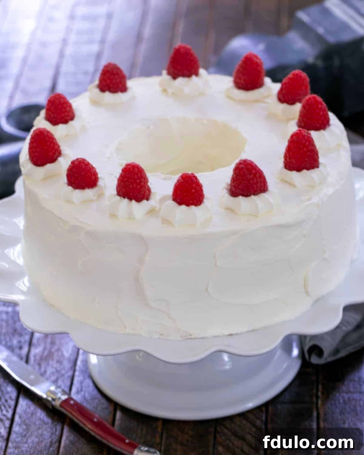 A beautifully frosted Angel Food Cake with its revealed chocolate mousse filling, elegantly garnished with fresh raspberries on a pristine white serving plate.