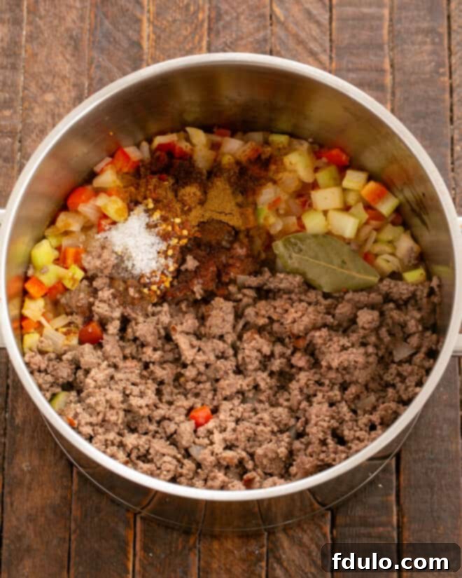 Close-up of ground beef browning in a pot with chili powder, cumin, and other seasonings, being broken apart with a spatula, indicating the spices are releasing their aromas into the meat.