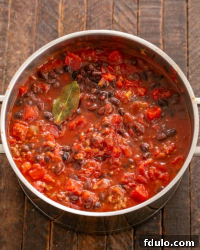 A large pot filled with all the chili ingredients—tomatoes, black beans, vegetables, and beef—simmering gently, with a bay leaf visible on the surface, indicating the flavors are melding.