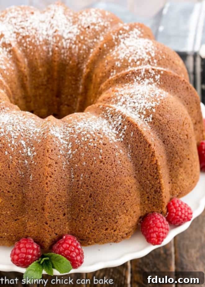Close-up view of a perfectly baked White Chocolate Bundt Cake on a cake stand, adorned with fresh raspberries and a sprig of mint.