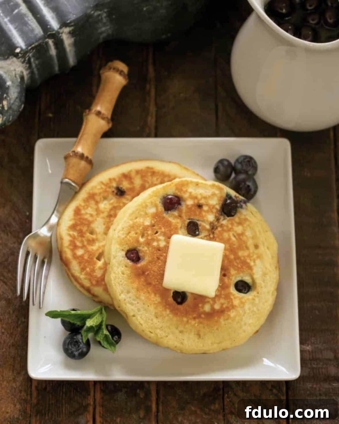 Two fluffy buttermilk pancakes with fresh blueberries on a square white plate, accompanied by a bamboo handle fork.