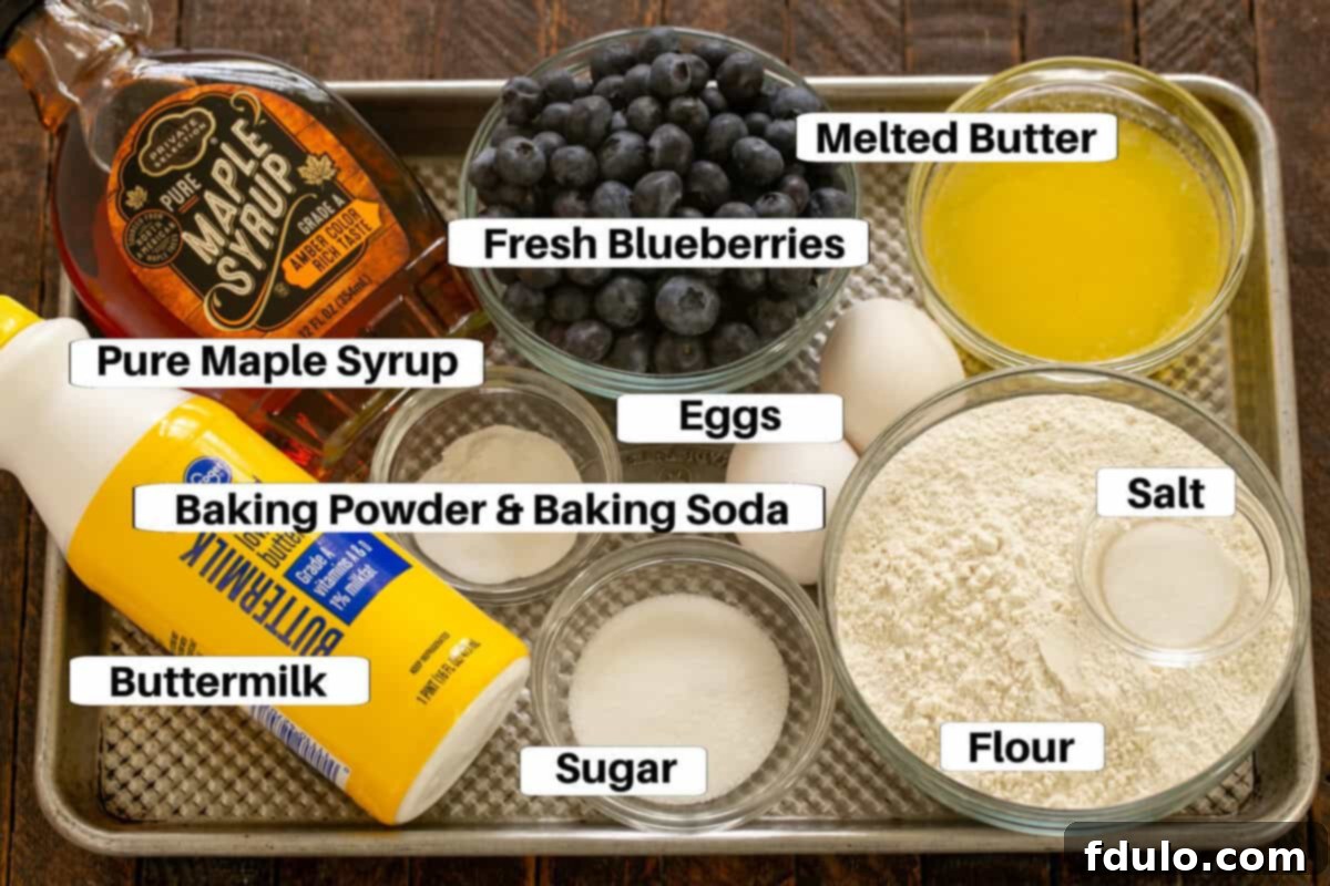 Pancake ingredients laid out on a metal sheet pan: flour, sugar, salt, baking soda, baking powder, eggs, buttermilk, melted butter, blueberries, and maple syrup.