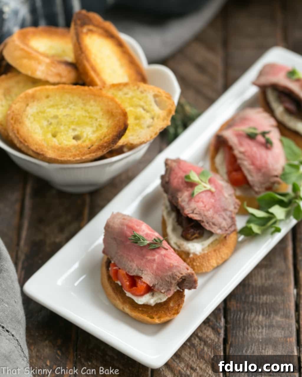Overhead view of Beef Crostini with Blue Cheese Spread and fresh herbs on a narrow white serving tray.
