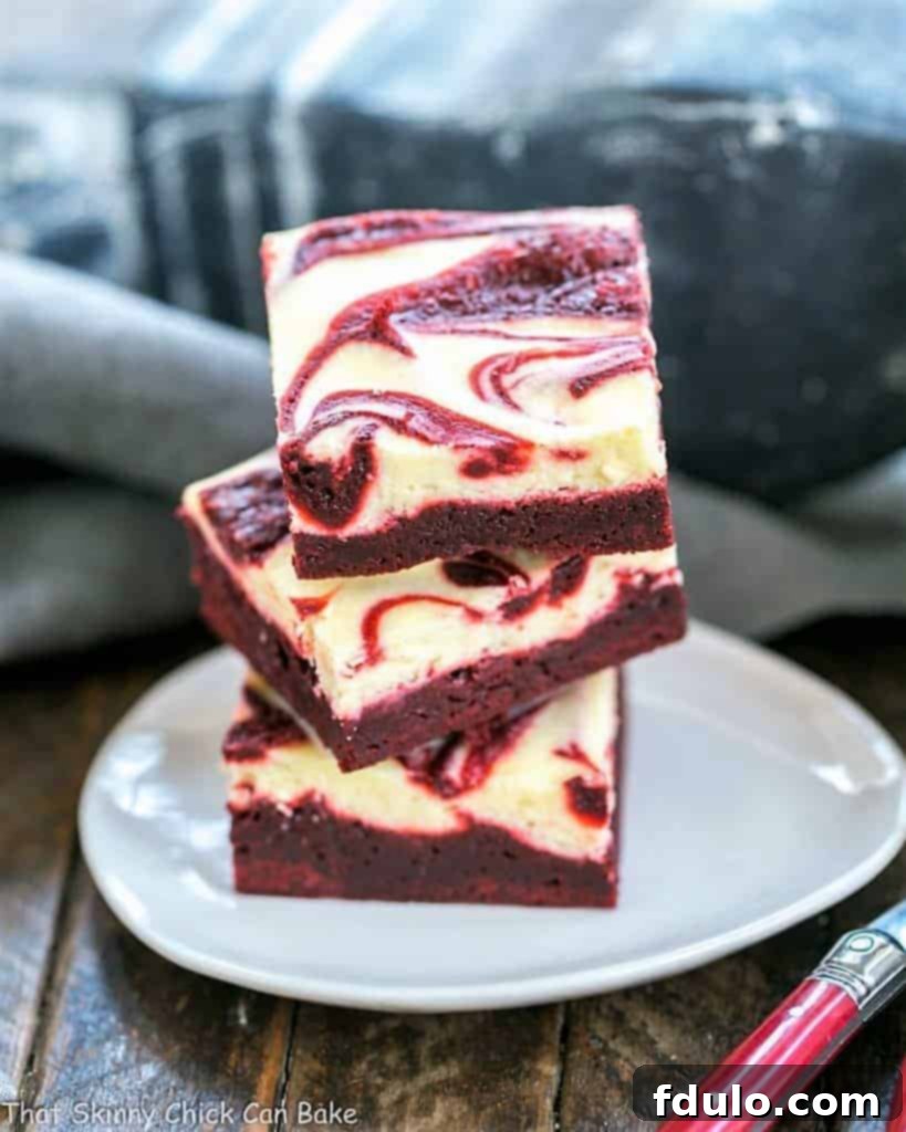 Three decadent swirled red velvet cheesecake brownies stacked on an elegant white tear drop shaped plate, showcasing their vibrant red and white layers.