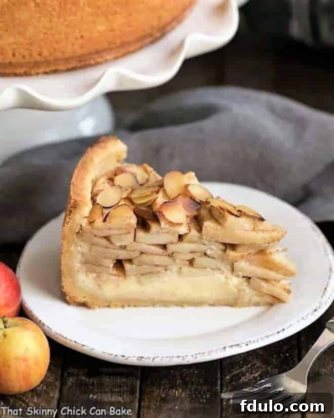 Cream Cheese Apple Torte on a white dessert plate with cake stand, apples and fork. A perfect blend of apple pie and cheesecake, garnished with fresh apples.