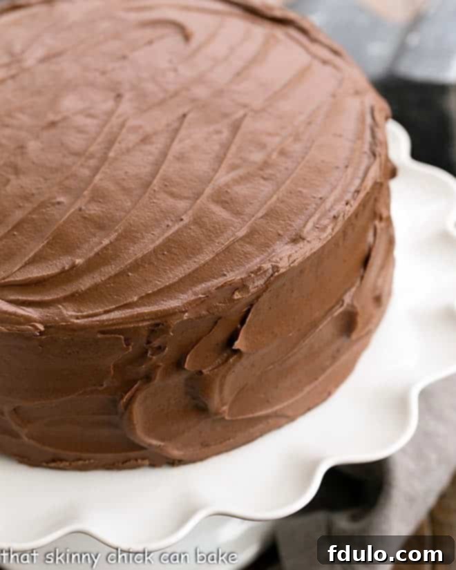 A stunning three-layer homemade chocolate cake elegantly presented on a white pedestal, covered in rich chocolate buttercream frosting, ready for serving.