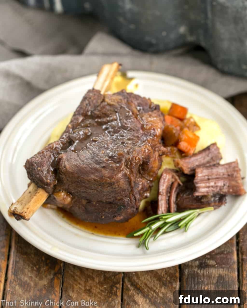 A gourmet serving of Instant Pot Beef Short Ribs, glistening with savory sauce and garnished with a fresh sprig of rosemary, presented on a sophisticated round white plate.