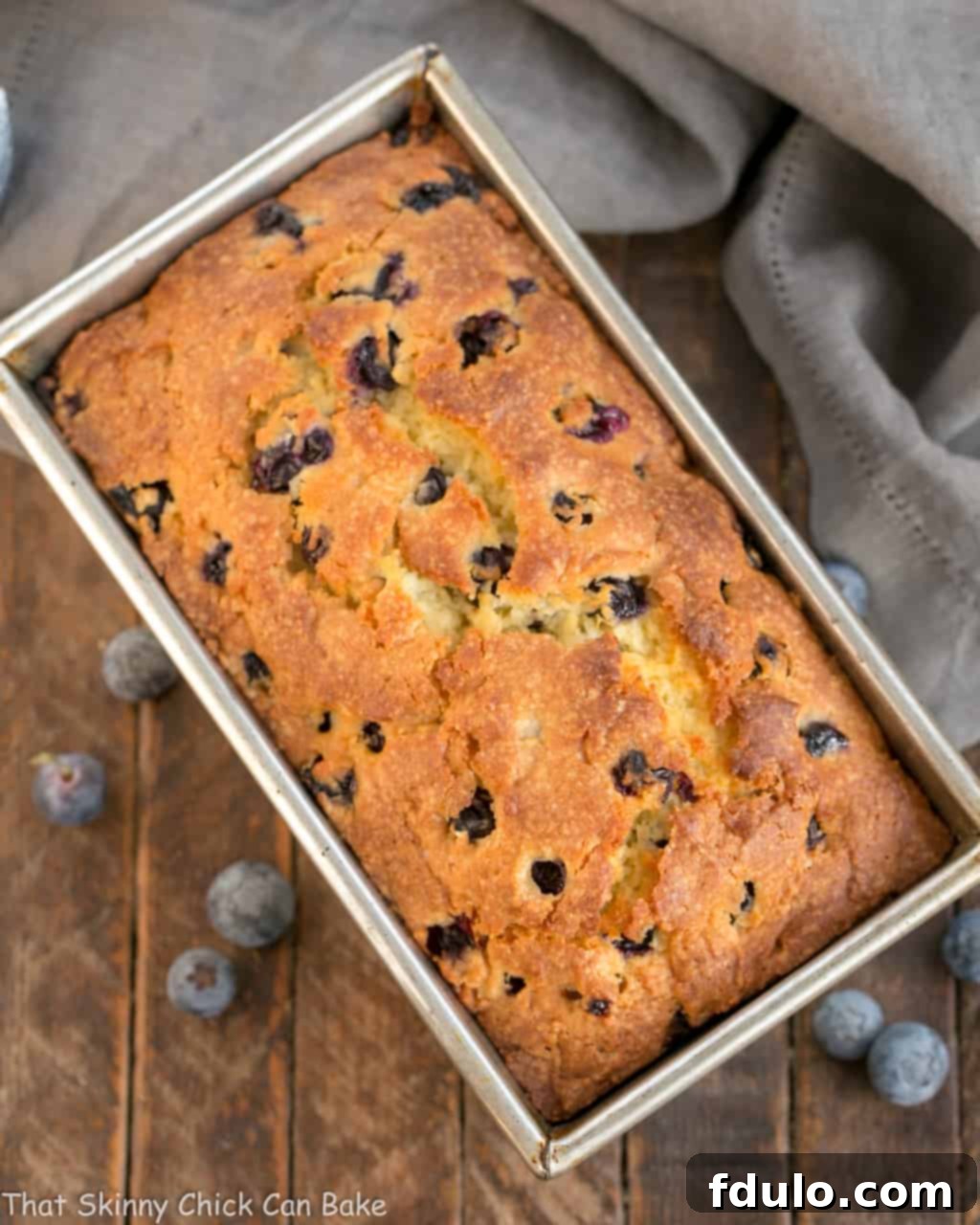Overhead view of a golden brown Lemon Blueberry Muffin Bread cooling in a loaf pan, brimming with blueberries.