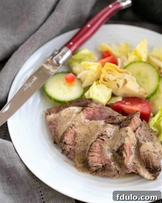 Bistrot Paul Bert Pepper Steak Recipe - perfectly pan-seared beef tenderloin with a luscious Cognac cream sauce Bistrot Paul Bert Pepper Steak sliced and drizzled with pan sauce with a fresh green salad on a white plate, showcasing its elegant presentation.