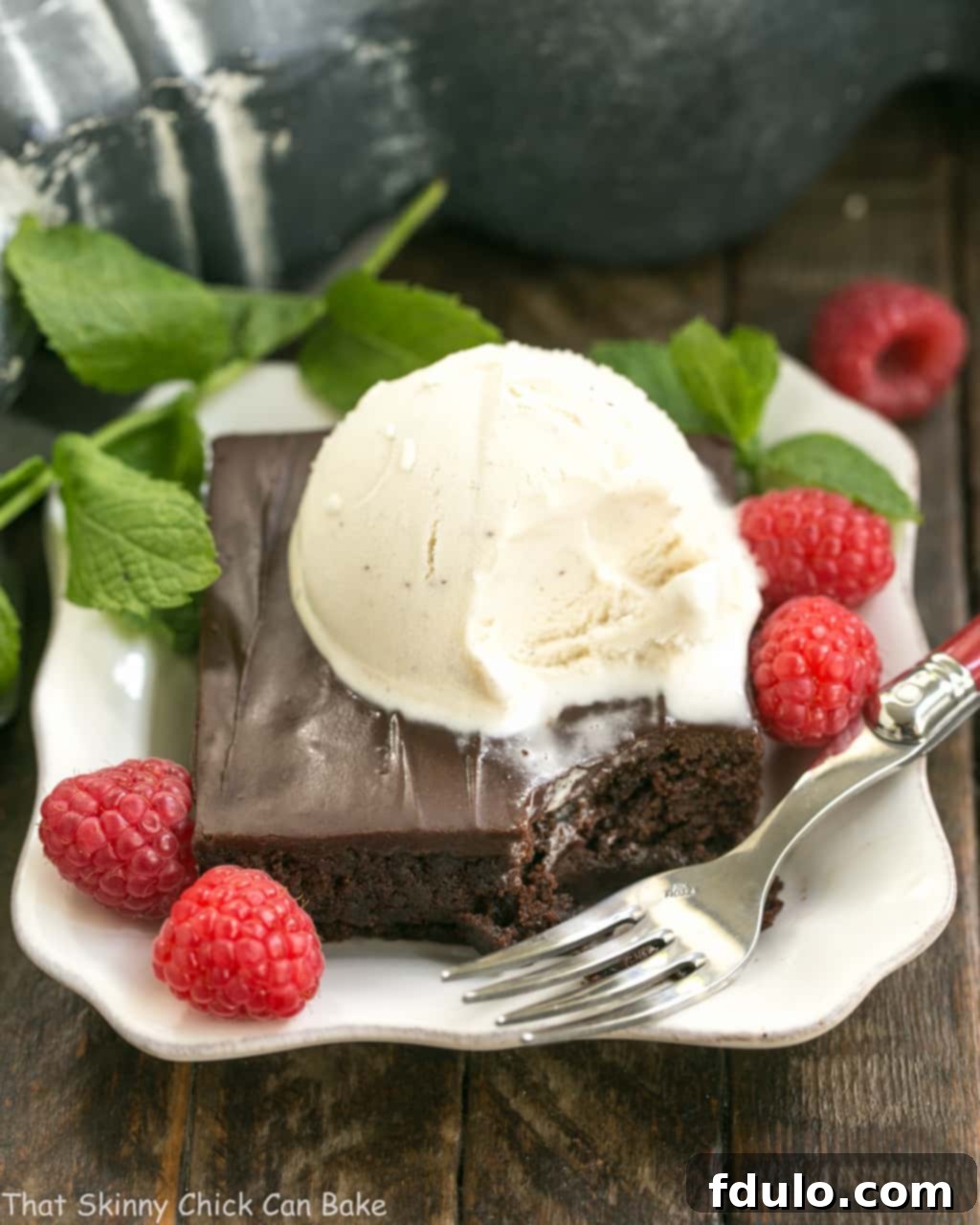 Fudgy Chocolate Sheet Cake with Cocoa Icing - A delectable, ultra-chocolate dessert that feeds a crowd! #sheetcake #chocolate A slice of fudgy chocolate sheet cake with rich cocoa icing, served on a white dessert plate alongside a scoop of vanilla ice cream, fresh mint leaves, and vibrant raspberries. This decadent dessert is perfect for any occasion.