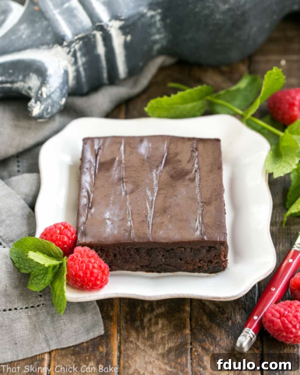 Fudgy Chocolate Sheet Cake with Cocoa Icing - A delectable, ultra-chocolate dessert that feeds a crowd! #sheetcake #chocolate A perfectly cut slice of Fudgy Chocolate Sheet Cake with smooth, shiny cocoa icing, presented on a minimalist square white plate and garnished with fresh raspberries and a sprig of mint for a pop of color.