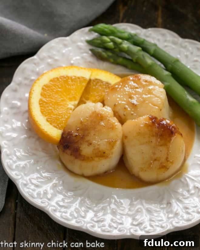 Pan seared scallops on a decorative white plate with orange slices and asparagus as garnish.