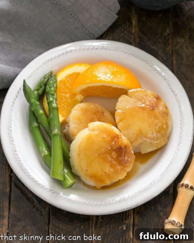Overhead view of pan seared scallops on a white plate with asparagus and fresh orange slices
