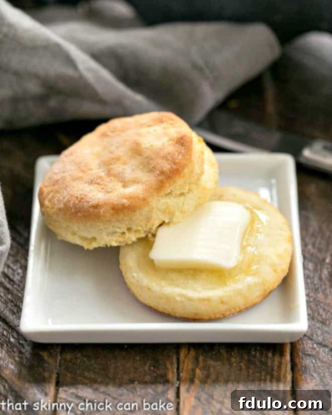 A freshly baked buttermilk biscuit, perfectly split to reveal its flaky layers, with a pat of melting butter nestled inside, presented on a square white plate.