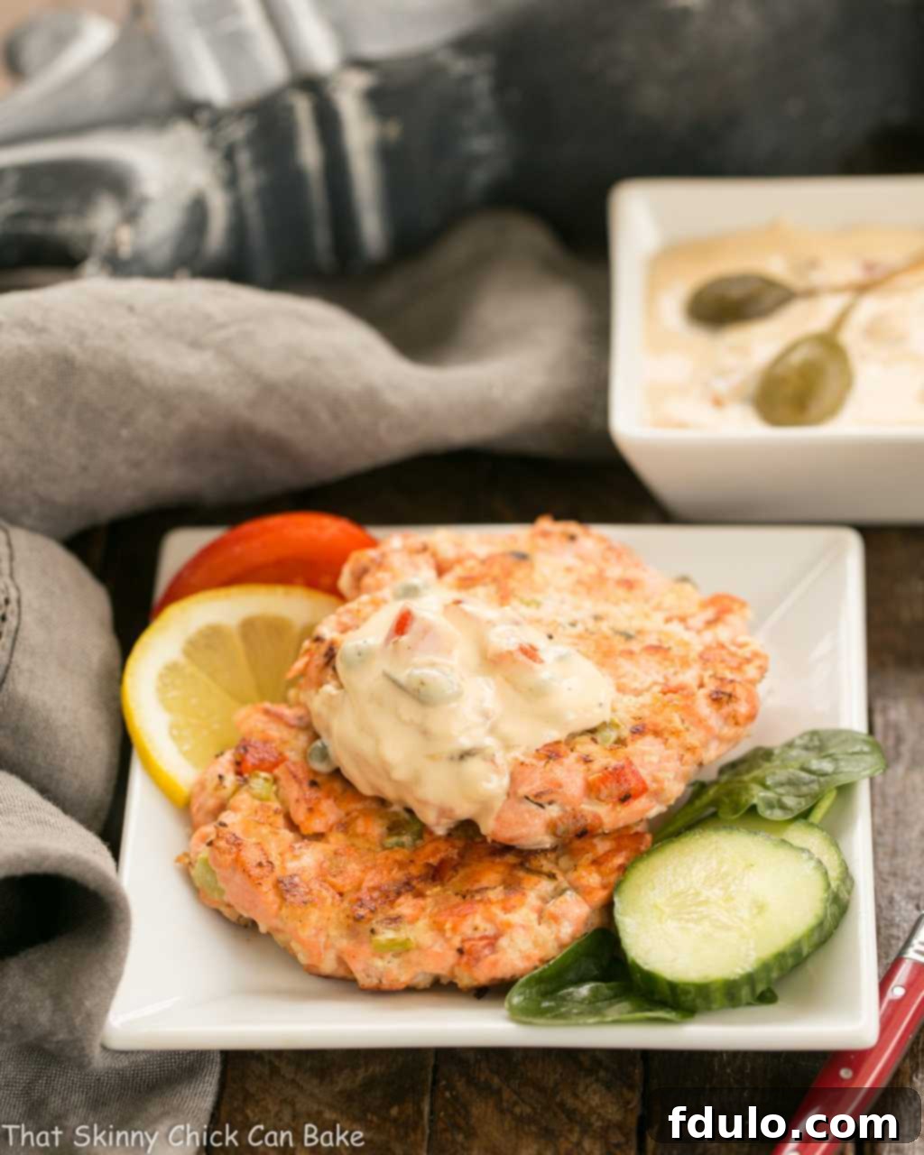 Two perfectly golden-brown fresh salmon cakes, beautifully garnished and arranged on a square white plate, ready to be served.