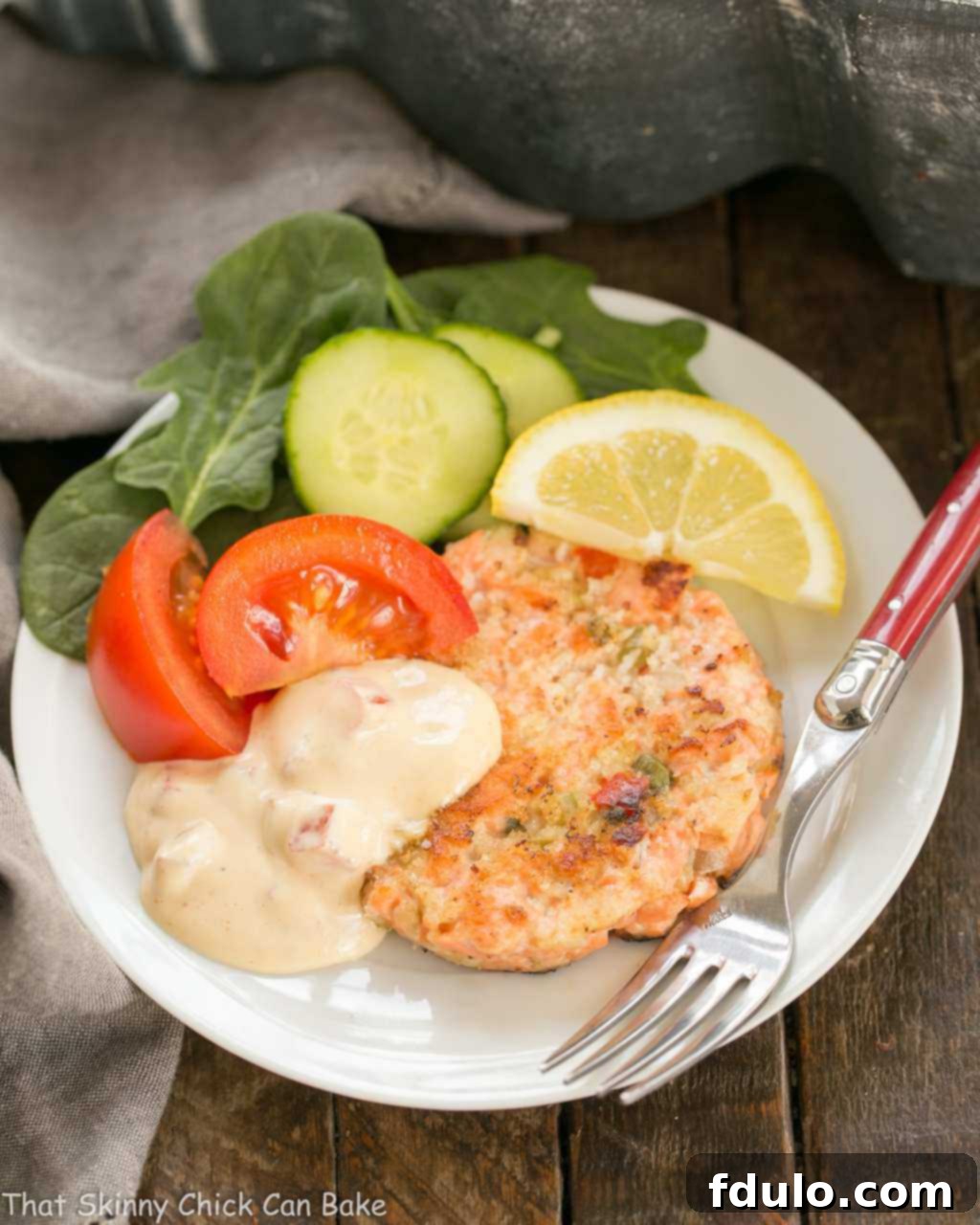 An overhead shot of a freshly cooked salmon cake, served with a vibrant green salad and a generous portion of creamy remoulade, all on a pristine white plate with a fork.