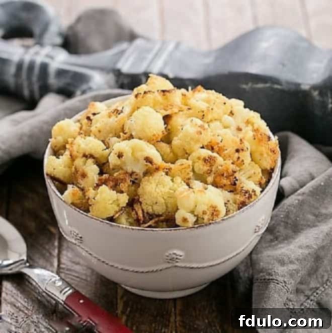 Golden-brown roasted cauliflower florets with visible Parmesan and breadcrumbs, served in a white ceramic bowl with a red-handled serving spoon, highlighting a delicious and satisfying side dish.